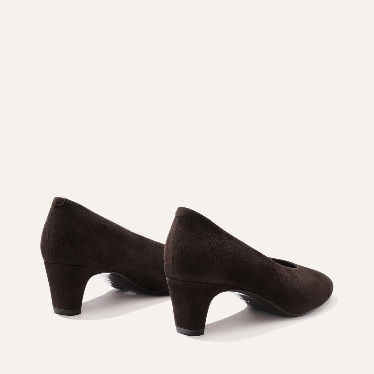 A pair of Margaux’s The Augusta Glove Pump in Molasses Suede, featuring a dark brown hue, rounded toes, and mid-height heels, are positioned side by side on a white background, angled slightly away.
