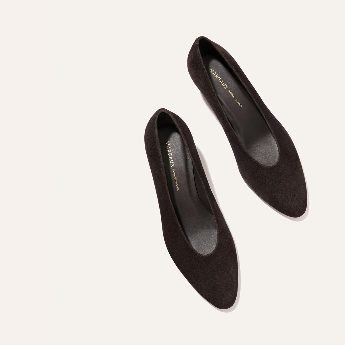 A pair of The Augusta Glove Pump in Molasses Suede by Margaux is displayed on a light beige background. The shoes are positioned diagonally, with the toes pointing to the upper right corner.