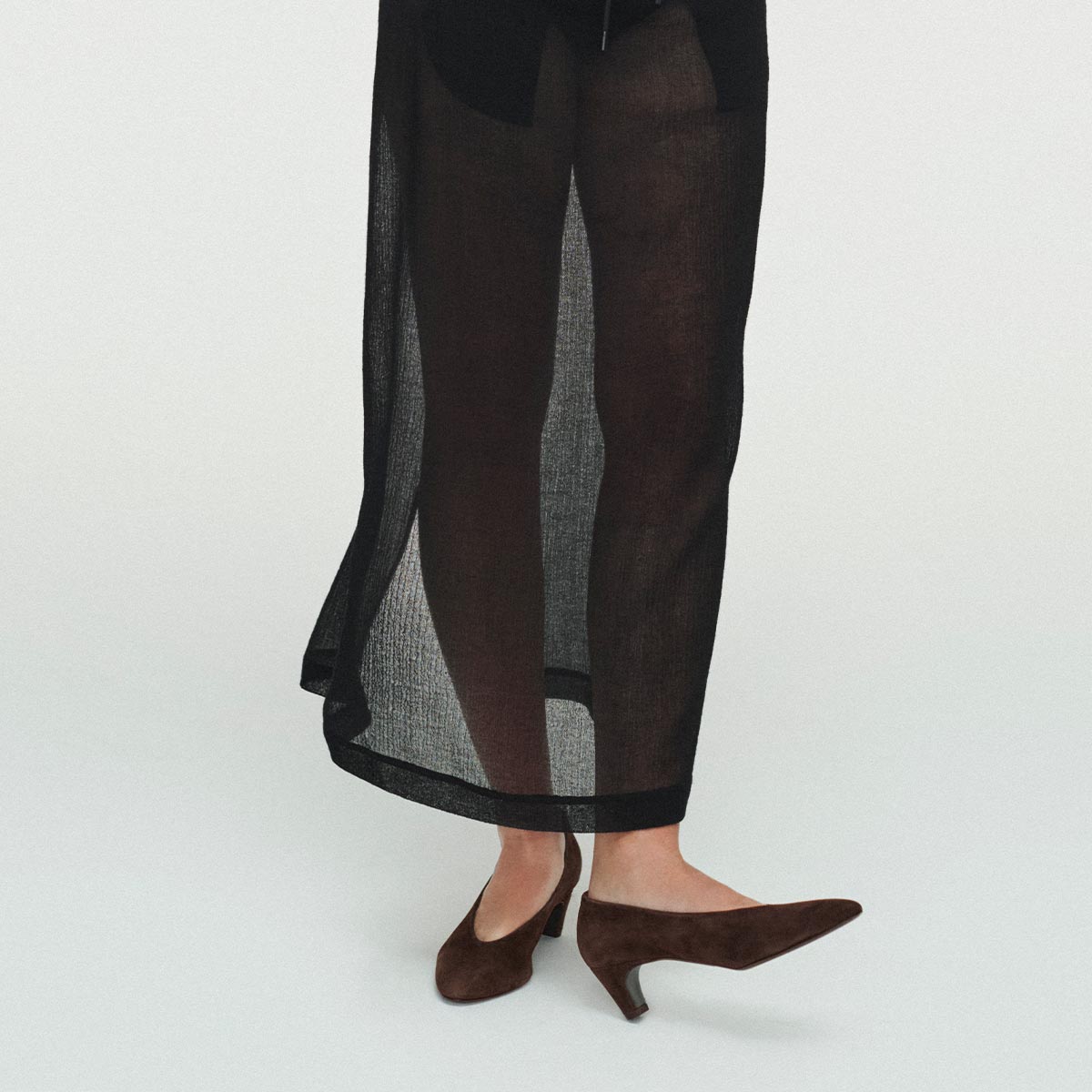 A person wears Margaux's The Augusta Glove Pump in Molasses Suede, styled with sheer black pants, standing against a white background. Only their legs and feet are visible in the image.