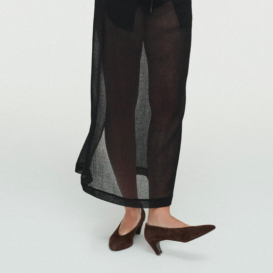 A person wears Margaux's The Augusta Glove Pump in Molasses Suede, styled with sheer black pants, standing against a white background. Only their legs and feet are visible in the image.