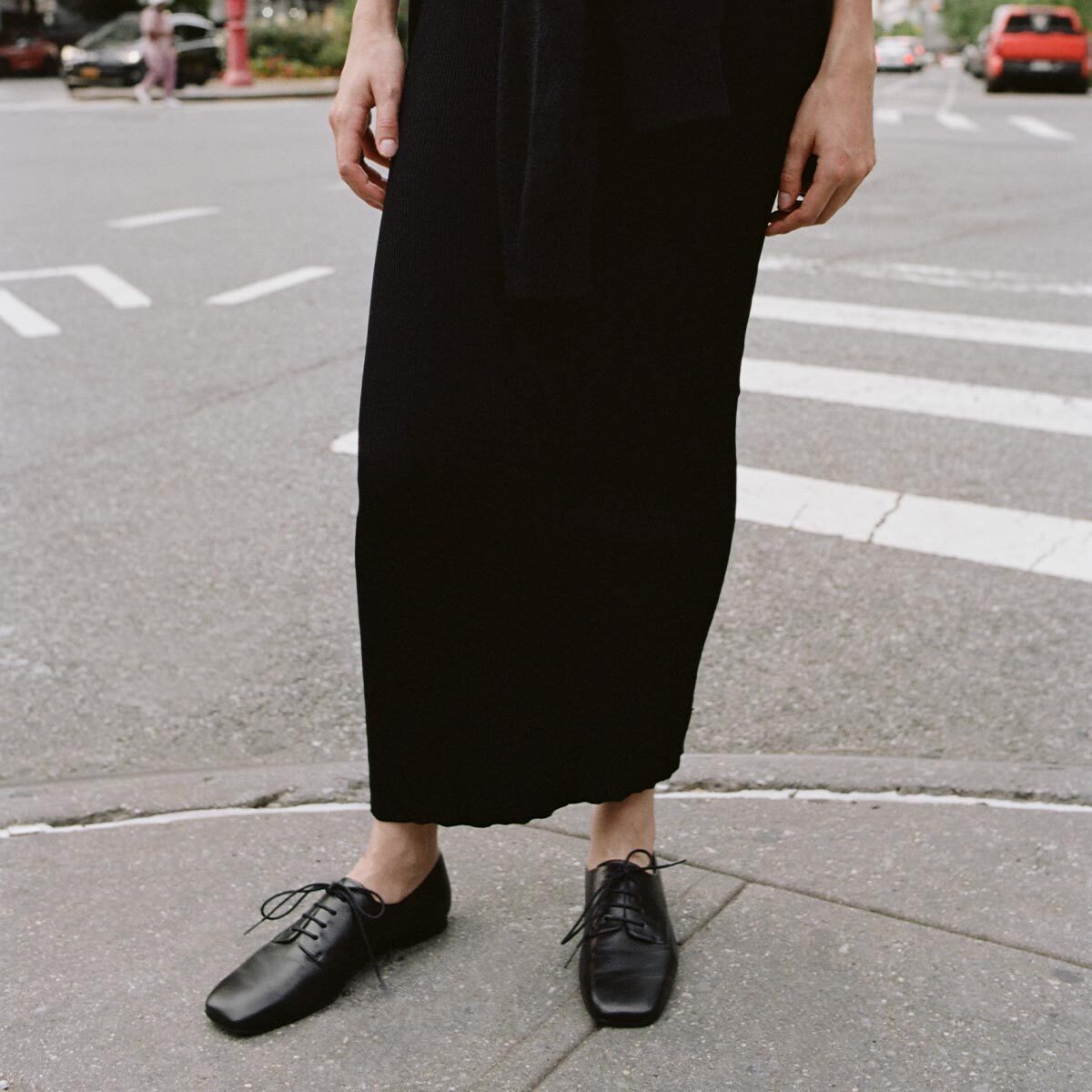 Standing on a city sidewalk, this person wears a long black skirt paired with Margaux’s The Billie Derby in Black Nappa, their hands relaxed at their sides as the street and crosswalk appear in the background.