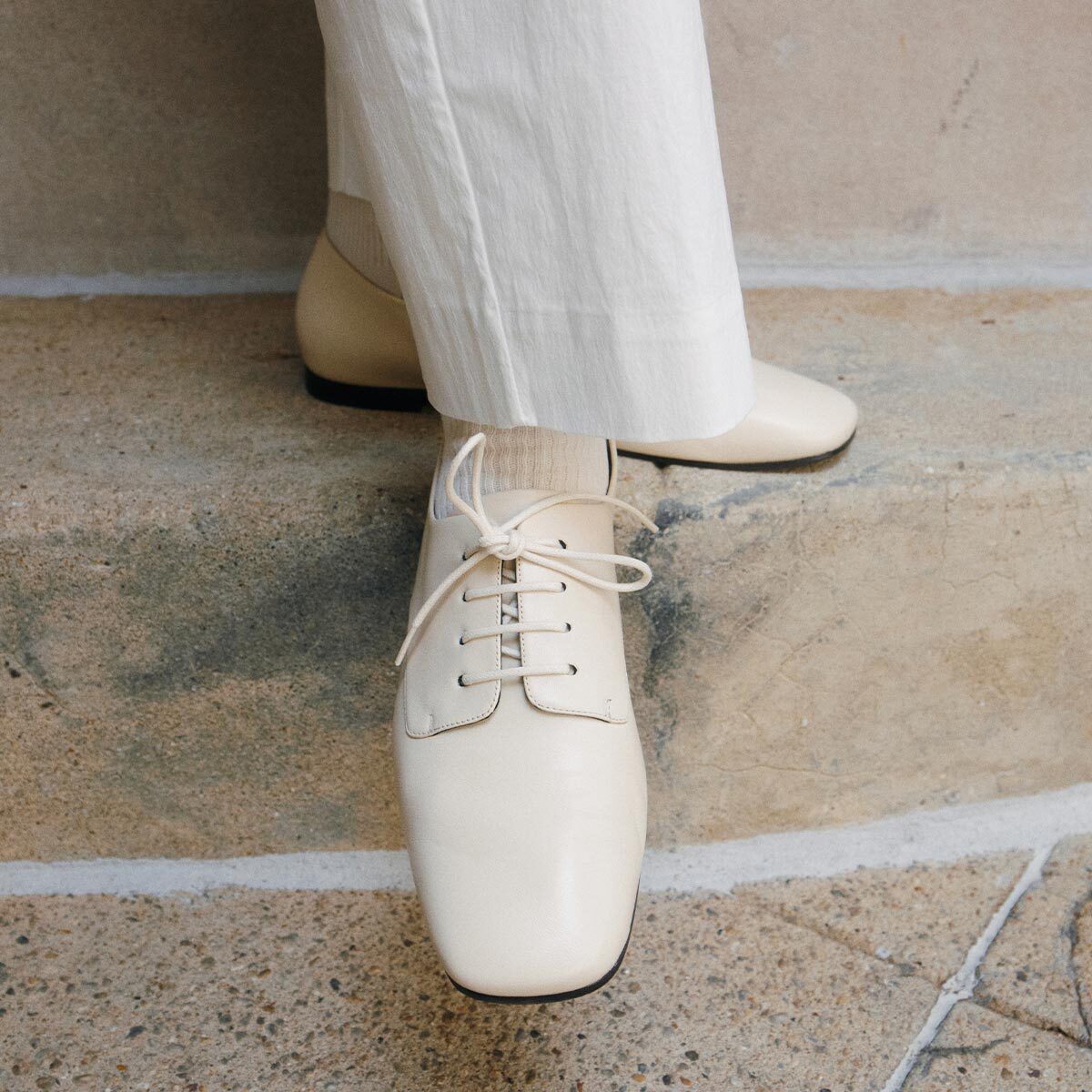 A person wearing cream-colored pants and Margaux’s The Billie Derby in Vanilla Nappa stands on stone steps, showcasing an effortless footwear choice.
