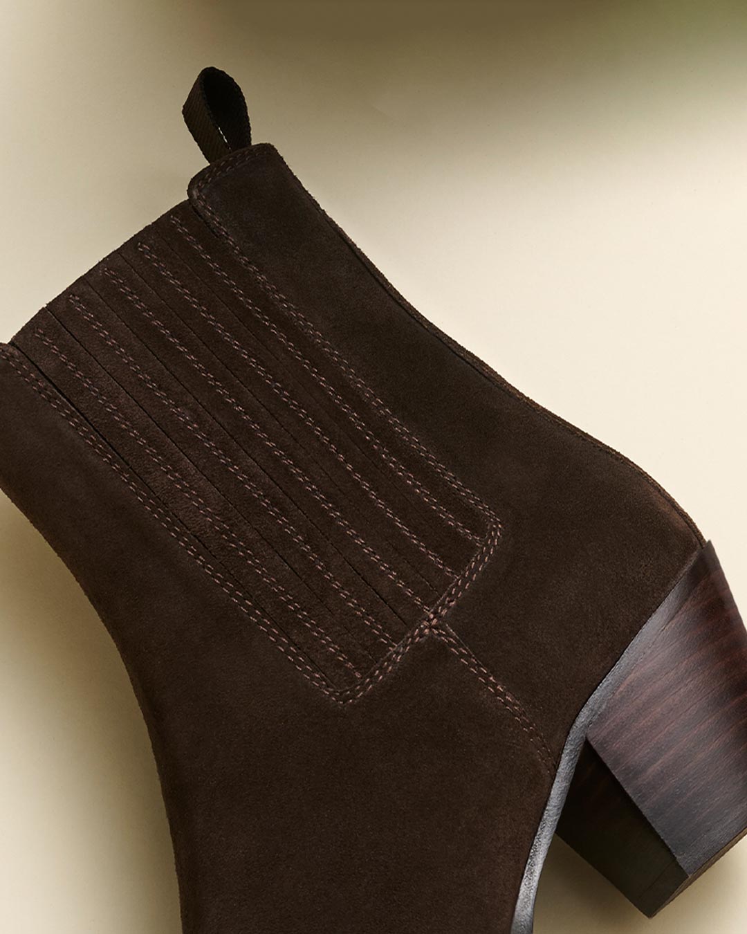A close-up of a dark brown suede ankle boot with vertical stitching details and a chunky, stacked wooden heel on a light beige background.
