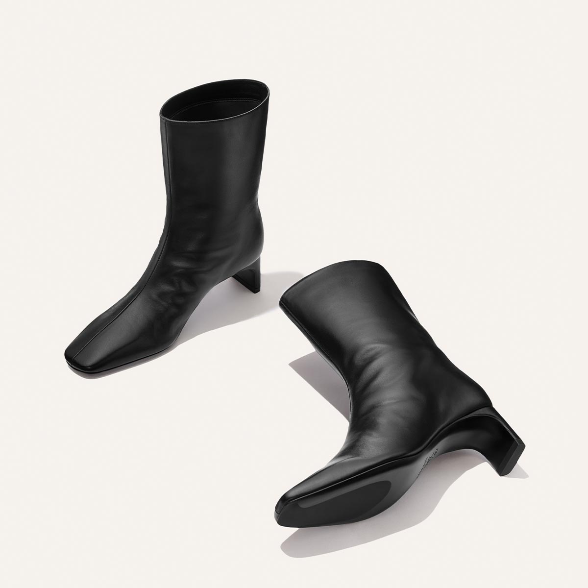 Two Brera Boots in black nappa leather by Margaux are displayed on a light background—one standing upright and the other lying on its side to reveal the sole. The mid-calf boots feature square toes and block heels.