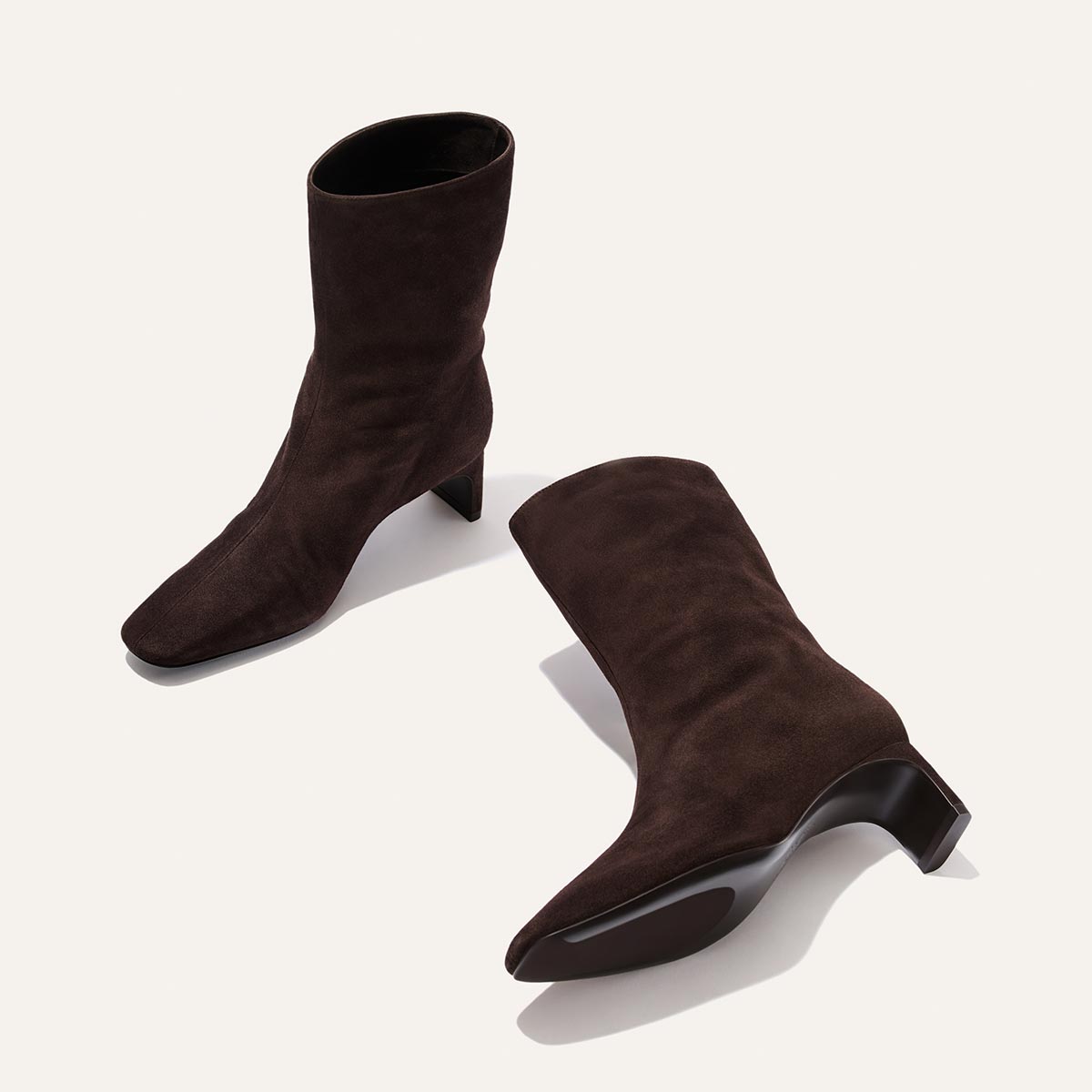 A pair of Margaux’s Brera Boot in Molasses Suede—mid-calf, with block heels—one standing upright and the other lying on its side, displayed against a light background.
