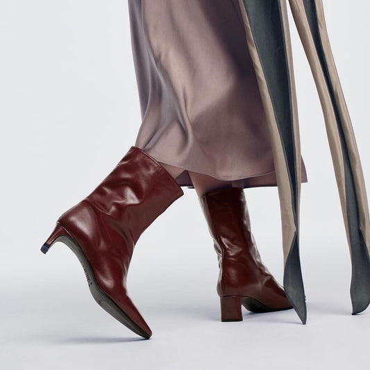 The Brera Boot - Merlot Calf, a pair of dark red leather mid-calf boots with square toes and 2″ heels, worn with a satin skirt.