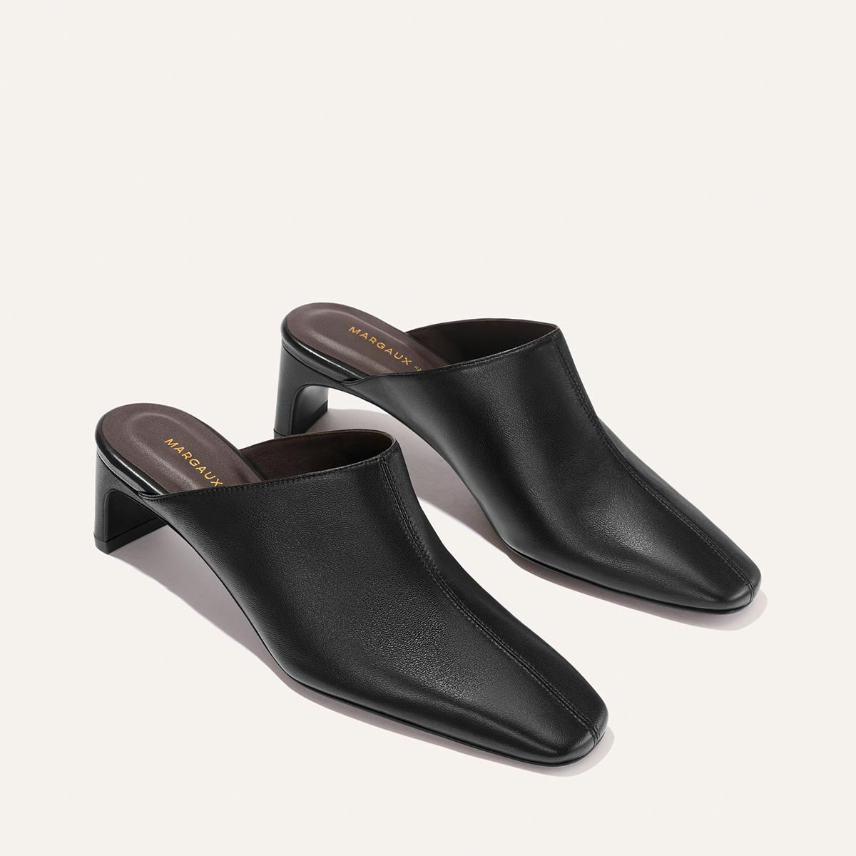 A pair of The Brera Mule - Black Nappa by Margaux, featuring black leather uppers with square toes and low block heels, displayed on a light beige background. Brown insoles with gold Margaux branding text are visible.