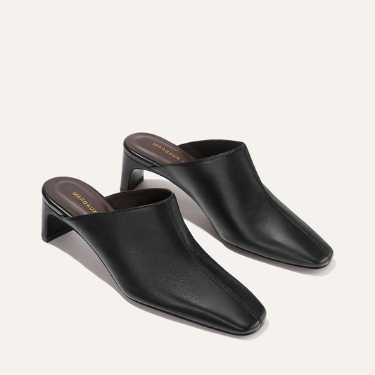 A pair of The Brera Mule - Black Nappa by Margaux, featuring black leather uppers with square toes and low block heels, displayed on a light beige background. Brown insoles with gold Margaux branding text are visible.