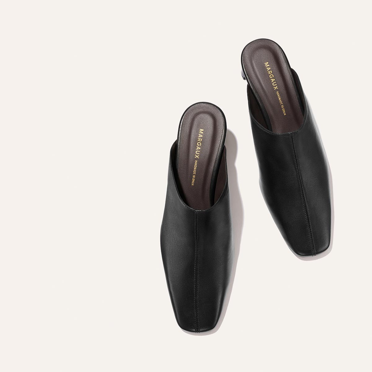 The Brera Mule - Black Nappa by Margaux, a pair of black leather slip-on mules with square toes, displayed from above on a light beige background. The dark brown insoles feature gold Margaux branding.