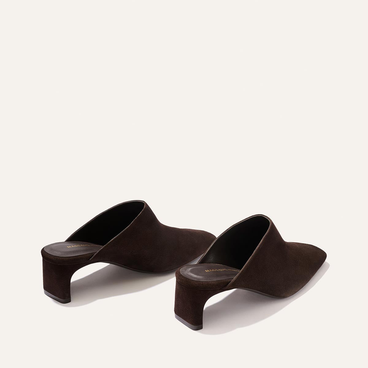 The Brera Mule - Molasses Suede by Margaux is an elegant pair of dark brown suede heeled mules featuring open backs, a high vamp, and curved block heels, displayed on a plain light background.