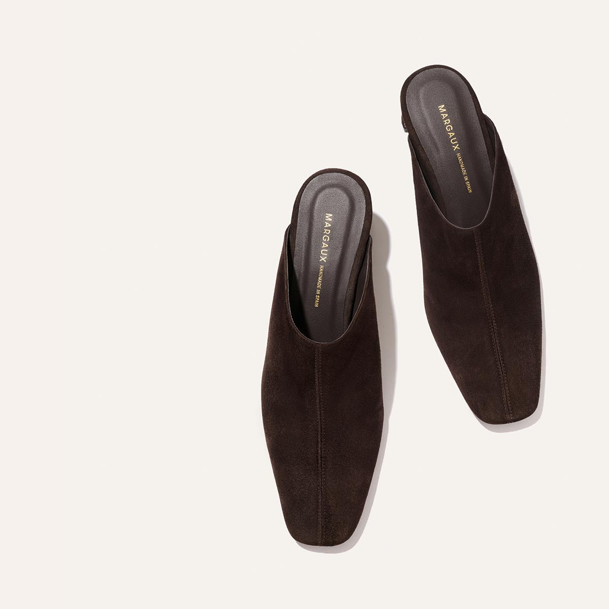 A pair of The Brera Mule in Molasses Suede by Margaux, featuring a dark brown suede slip-on design with square toes and a high vamp, viewed from above on a light background. The dark insoles display gold-printed Margaux branding.