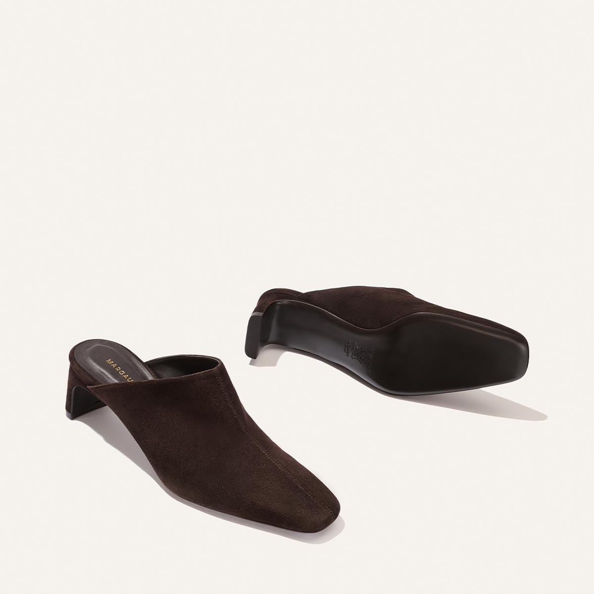 The Brera Mule by Margaux in Molasses Suede is displayed on a plain light background, showcasing its dark brown suede upper, high vamp, and square toe. The elegant silhouette is complemented by a low sculpted heel, with one shoe positioned upright and the other revealing the sole.