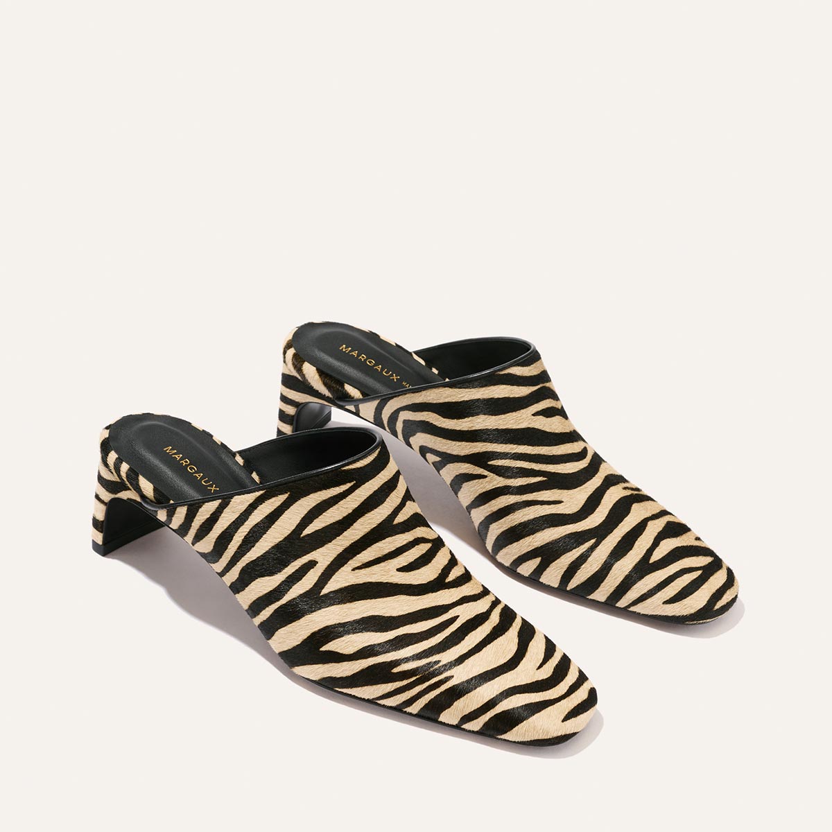 A pair of The Brera Mule - Tan Zebra Haircalf by Margaux, featuring a black and beige zebra print pattern and black insoles, displayed on a light background.