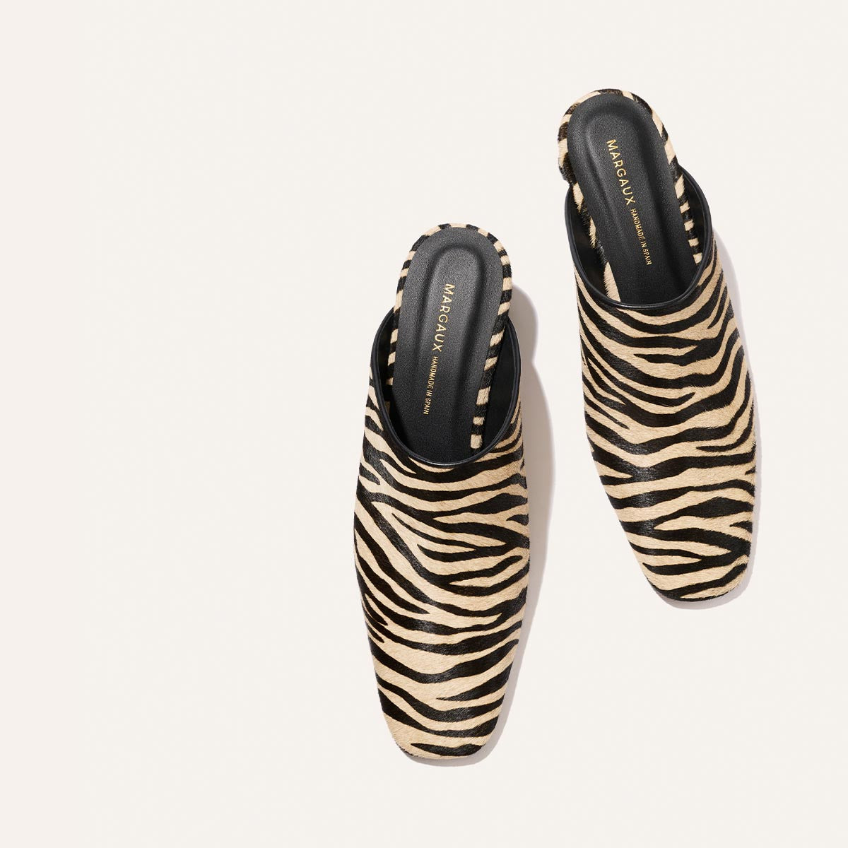 A pair of Margaux’s The Brera Mule in Tan Zebra Haircalf, featuring black insoles, positioned diagonally on a light, neutral background.