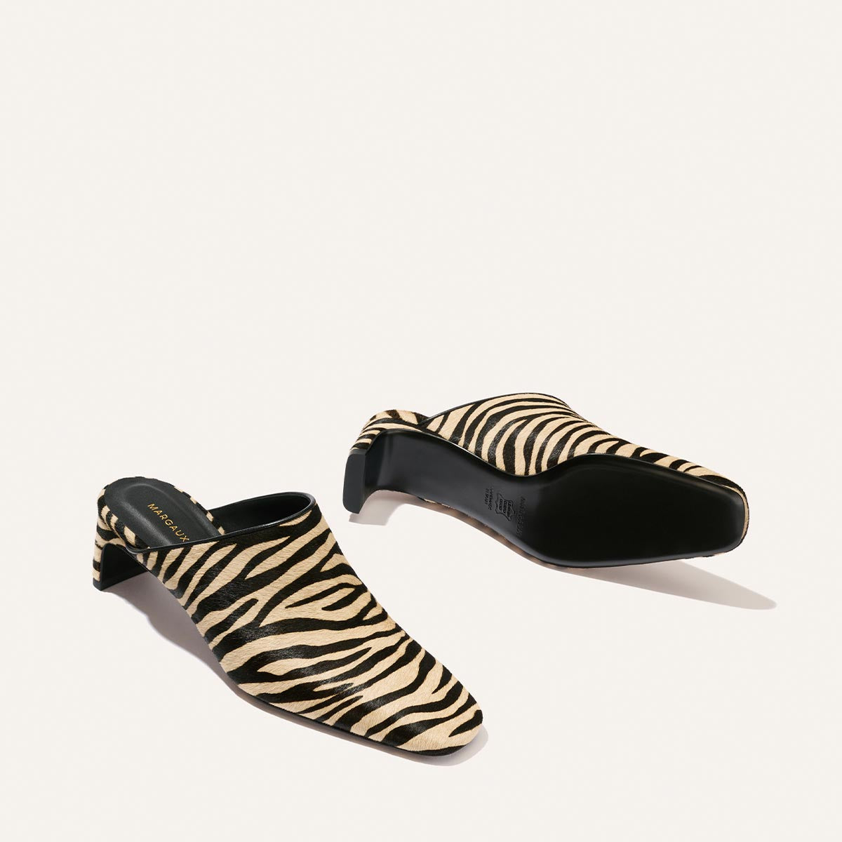 A pair of Margaux’s The Brera Mule in Tan Zebra Haircalf, featuring a black and beige zebra print pattern—one shoe displayed upright while the other lies on its side to showcase the sole.