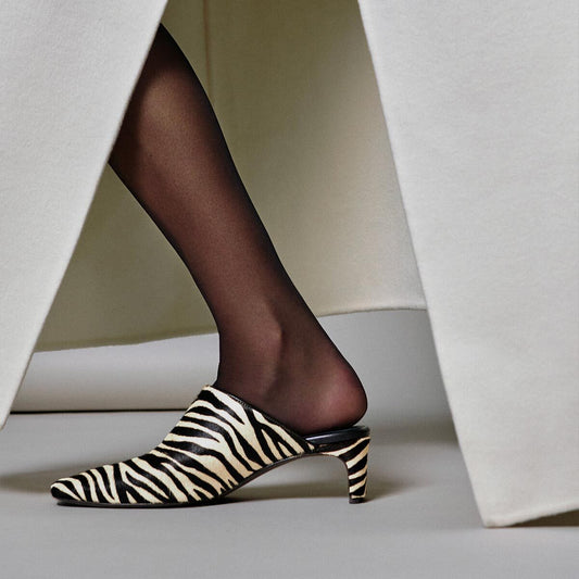A person wearing black sheer stockings and Margaux’s The Brera Mule in Tan Zebra Haircalf steps forward between two white fabric panels.