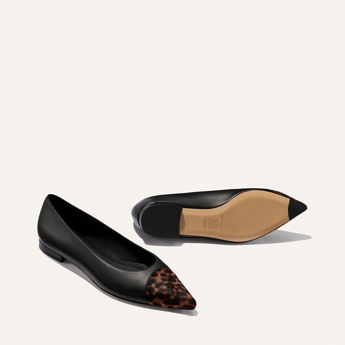 Two "The Alba" flats by Margaux in black nappa leather feature a brown leopard haircalf detail on the pointed toe. One shoe is upright, while the other lies on its side, displaying the beige sole and suggesting a comfortable, padded insole.