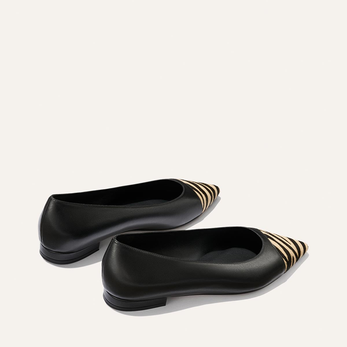 A pair of The Alba ballet flats by Margaux, crafted from black nappa leather and featuring tan and black zebra haircalf detailing on the pointed toes, with a padded insole for extra comfort, shown against a plain white background.