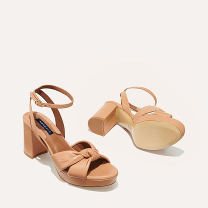 The Carmine Platform - Desert Nappa by margaux-development are tan high-heeled sandals featuring ankle straps, chunky platform heels, and plush knot detailing. One shoe stands upright while the other lies on its side, displaying the sole.