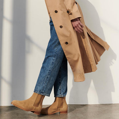 A person wearing a beige trench coat, blue jeans, and Margaux’s The Chelsea Boot in Sand Suede walks indoors. The image, cropped to the lower body and feet, shows a soft shadow cast on a white wall.