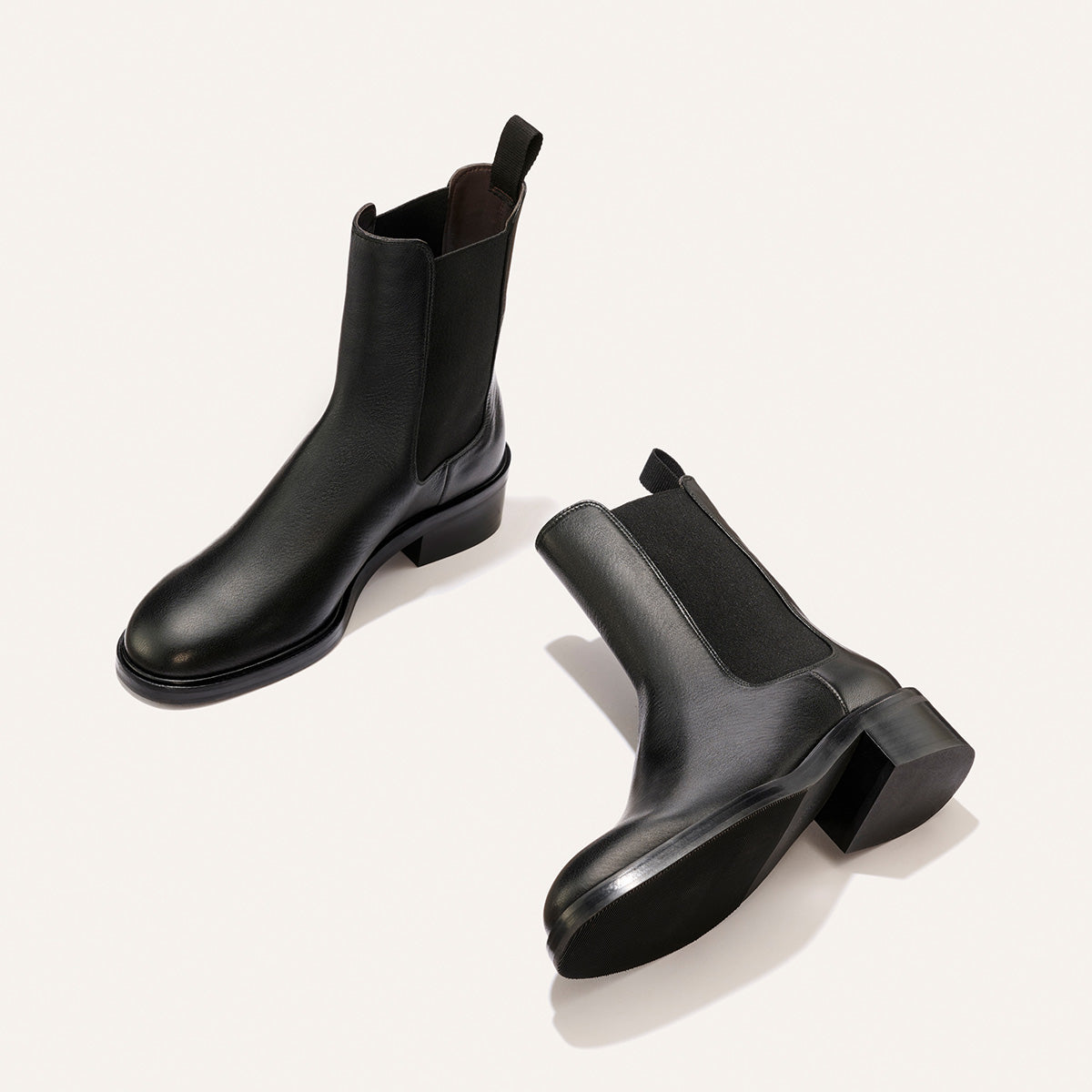 A pair of The City Chelsea Boot - Black Calf by Margaux, crafted from black leather with elastic side panels and pull tabs, is displayed on a light, neutral background. One boot stands upright while the other lies on its side to reveal the sole.