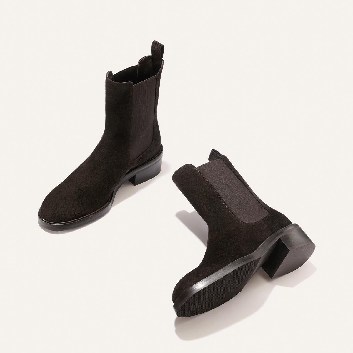 A pair of Margaux’s The City Chelsea Boot in Dark Chocolate Suede, one standing upright and the other lying on its side, both featuring elastic side panels and pull tabs, displayed on a light background.