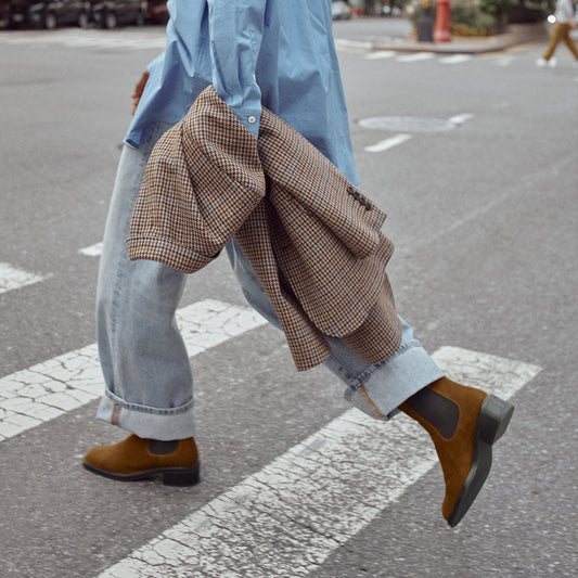 A person crossing the street in cuffed jeans shows movement, their lower body visible as they wear Margaux’s The City Chelsea Boot in Cedar Suede.