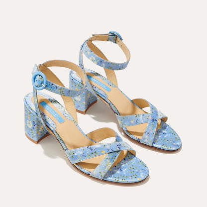 The City Sandal - Blue Floral Satin by margaux-development features a light blue floral print, crossed toe straps, ankle straps with round blue buckles, block heels, and a cushioned insole for extra comfort.