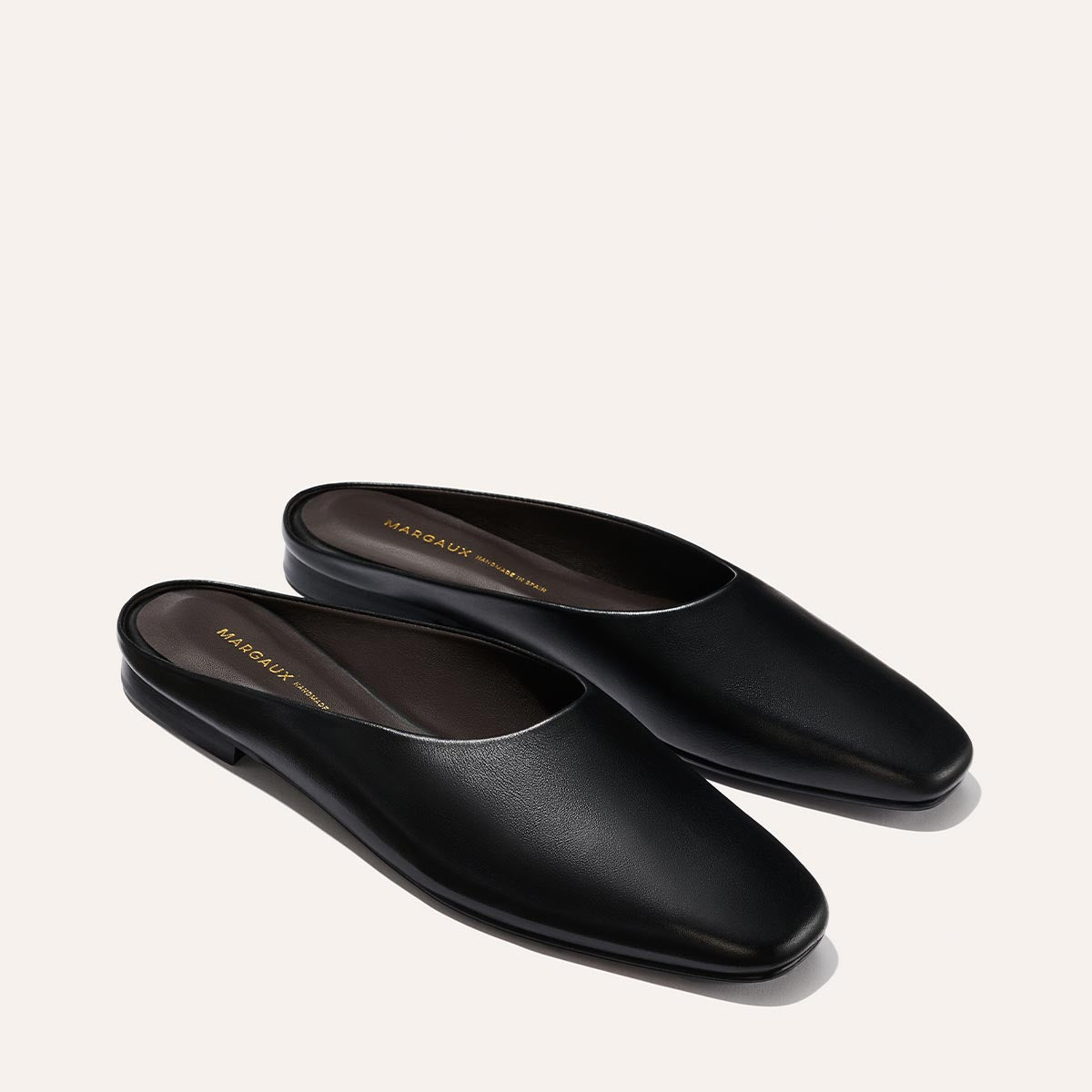 A pair of Margaux The Clara - Black Calf leather slip-on mule shoes with a closed, rounded square toe and flat sole is displayed on a light background, featuring dark interior lining and gold brand lettering inside.