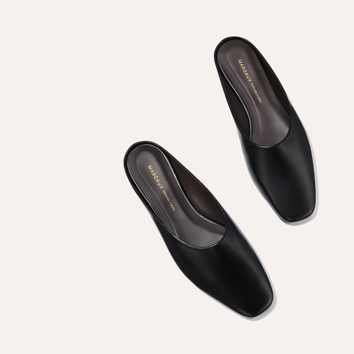 A pair of The Clara - Black Calf slip-on mules by Margaux, featuring black leather and square toes, are placed diagonally on a light beige background. The insole displays the brand’s gold logo.