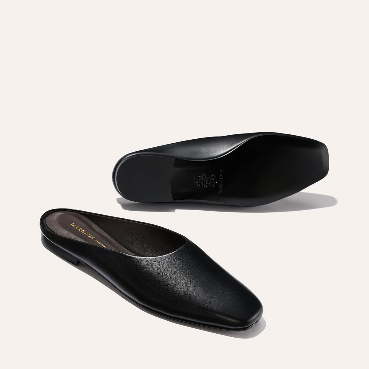 A pair of Margaux The Clara - Black Calf leather mules, one shown in side view and the other displaying the sole, set against a light background.