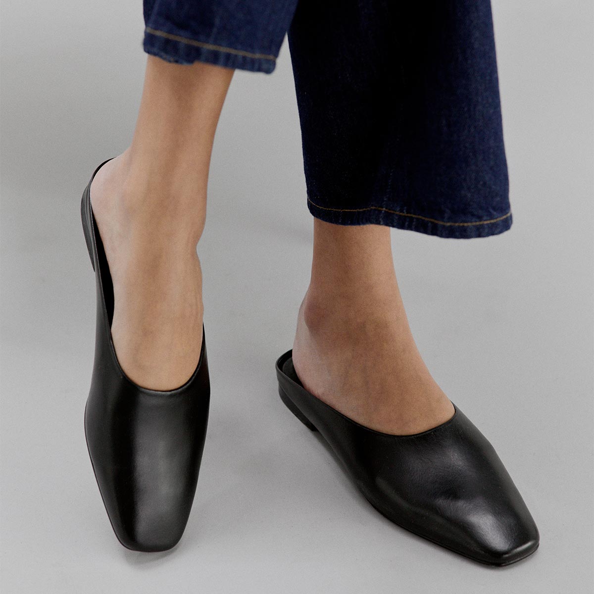 Wearing dark blue jeans, a person stands on a light gray surface in Margaux’s The Clara - Black Calf slip-on mules, highlighting their versatile, modern slipper silhouette.