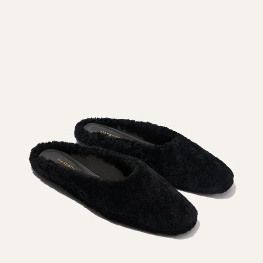 A pair of The Clara - Black Shearling slippers by Margaux, featuring a black fuzzy shearling finish with closed toes, rest on a plain off-white background. These slippers showcase a flat silhouette and plush texture for soft, cozy indoor comfort and versatile style.