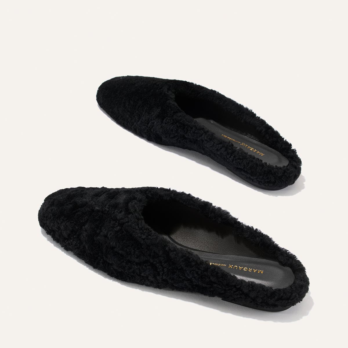 A pair of black shearling backless slippers, The Clara by Margaux, featuring a flat slipper-like silhouette and displayed on a plain light background.
