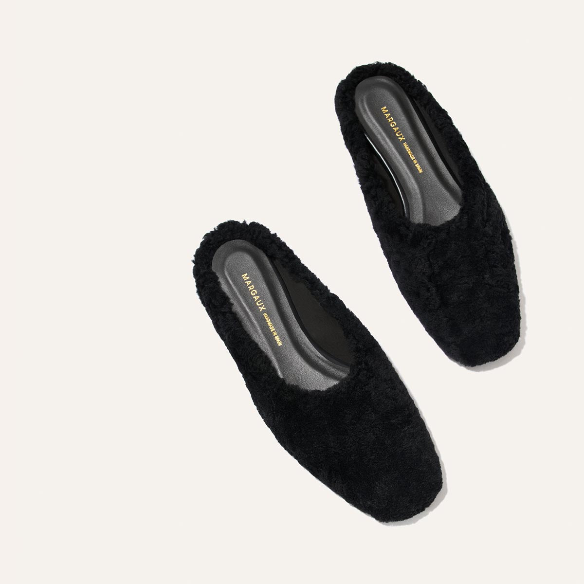 A pair of The Clara - Black Shearling slippers by Margaux, featuring a black fuzzy slip-on design with a slight heel and versatile style, are placed on a light background. The insole displays MARGAUX NEW YORK in gold lettering.
