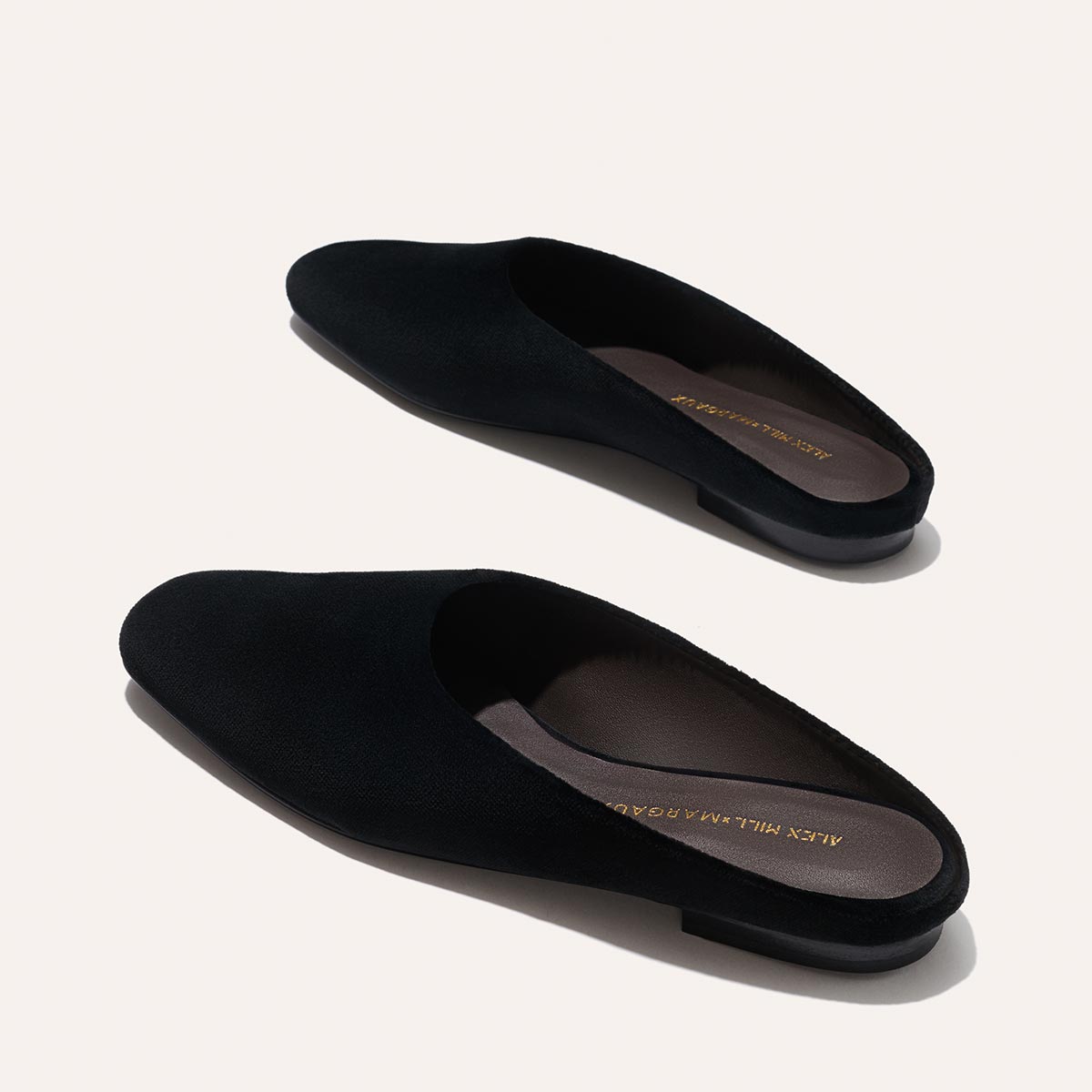 The Clara - Black Velvet by Margaux are black, slip-on, backless loafers with low heels, shown on a light background. These flats feature gold branding on the insoles and offer stylish versatility.