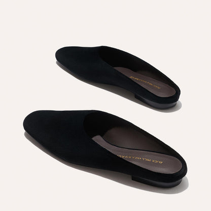 The Clara - Black Velvet by Margaux are black, slip-on, backless loafers with low heels, shown on a light background. These flats feature gold branding on the insoles and offer stylish versatility.