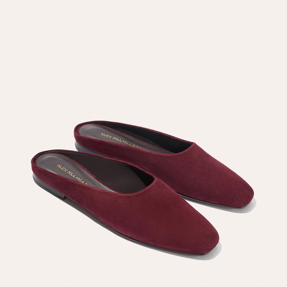 The Clara by Margaux for Alex Mill is a pair of burgundy suede slip-on mules with flat black soles and a closed-toe design, shown on a light background—an elegant, versatile choice for any occasion.