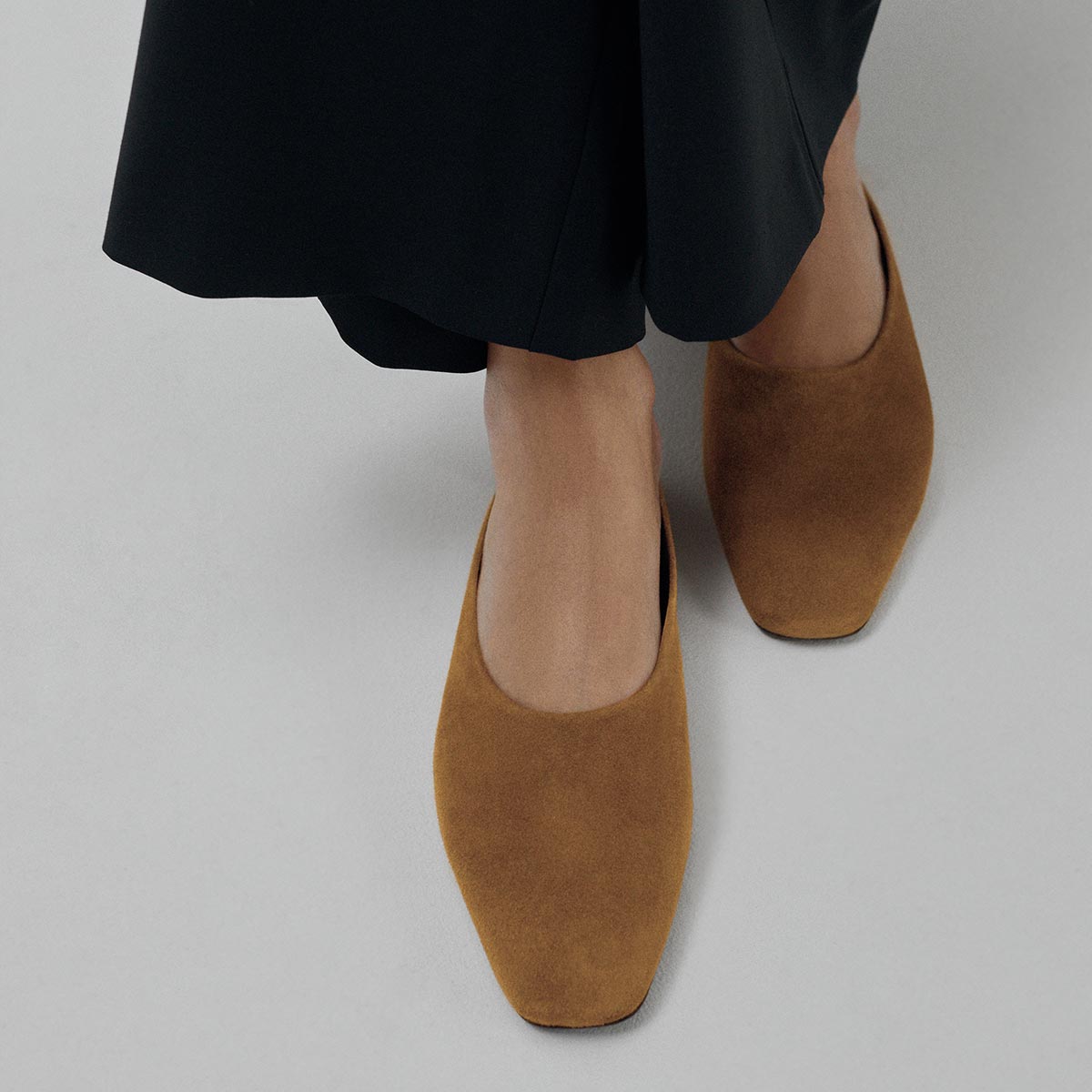 A person wearing black pants and Margaux's The Clara - Cedar Suede flats with a versatile style and slipper-like silhouette, standing on a light gray surface.
