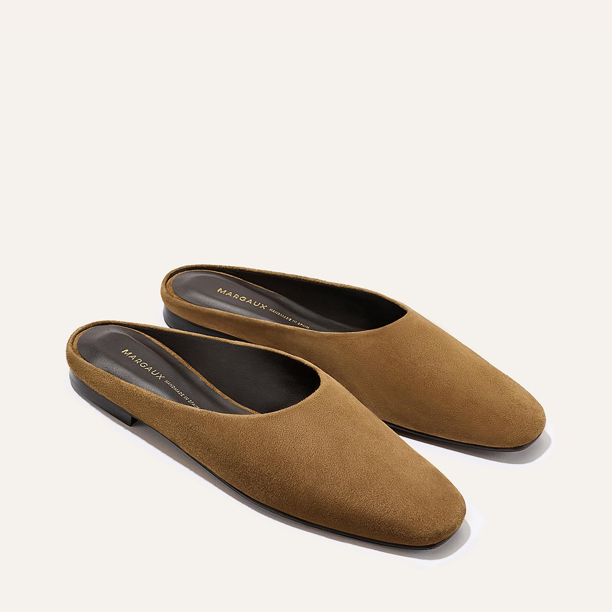 A pair of The Clara - Cedar Suede slip-on flats by Margaux, featuring brown suede, closed rounded toes, low heels, and branded insoles, displayed against a light background.