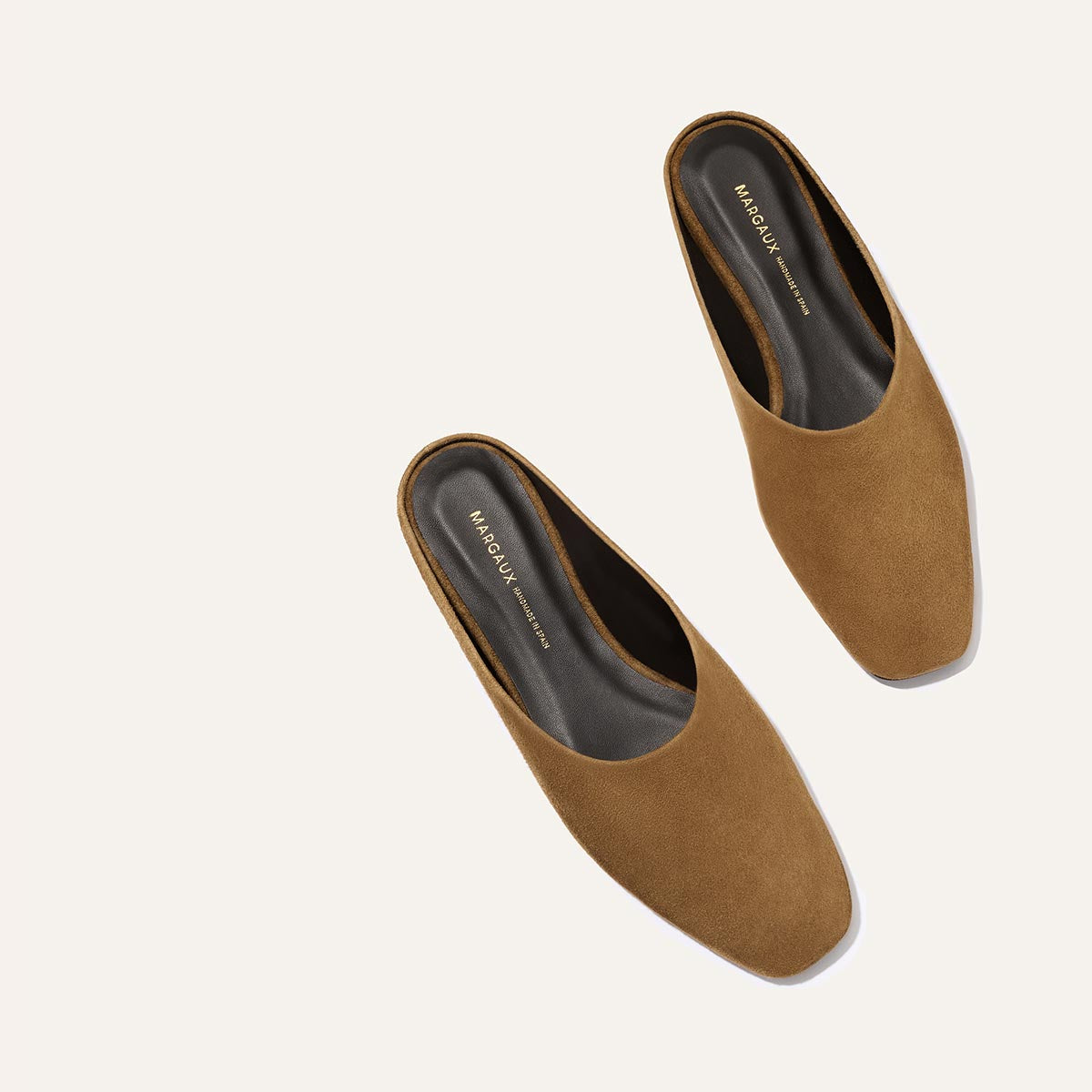 The Clara - Cedar Suede by Margaux: brown suede mules with black insoles, styled diagonally on a light background. These shoes feature a minimalist look with closed, gently squared toes.