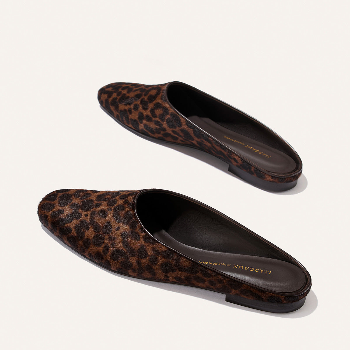 Two slip-on mules, The Clara in Chocolate Leopard Haircalf by Margaux, are displayed on a plain white background. These shoes feature a closed toe and open back with a flat sole, delivering versatile style and effortless, slipper-inspired comfort for all-day wear.