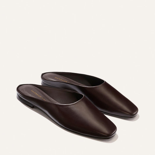 Introducing The Clara - Coffee Calf by Margaux: a pair of dark brown leather slip-on mules with a flat, versatile design and closed, rounded toes. Featuring a slipper-like silhouette, these shoes are beautifully displayed on a light background.