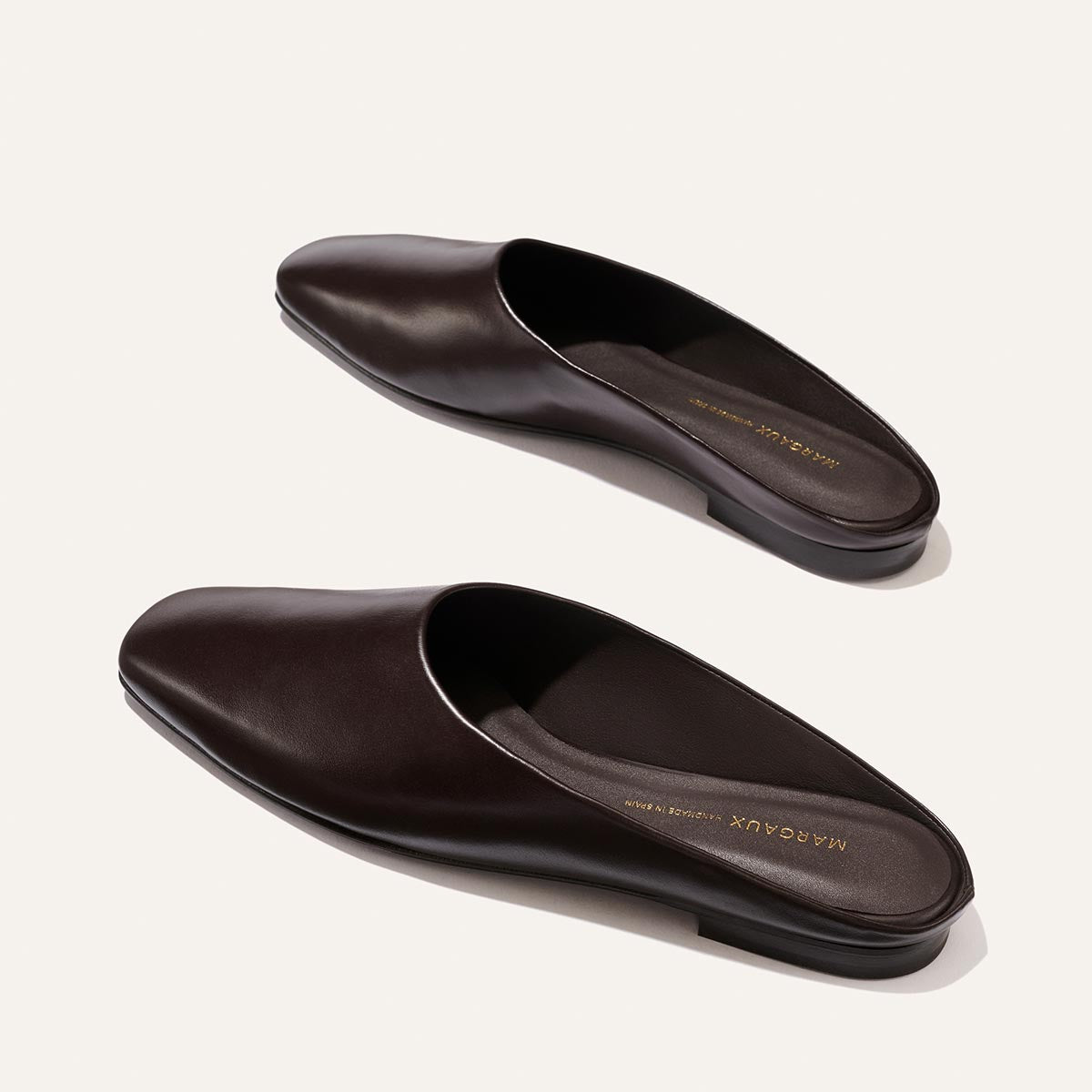 Two coffee-colored leather slip-on mules, The Clara by Margaux, featuring a slipper-like silhouette and flat soles, are placed on a light background, oriented in opposite directions.