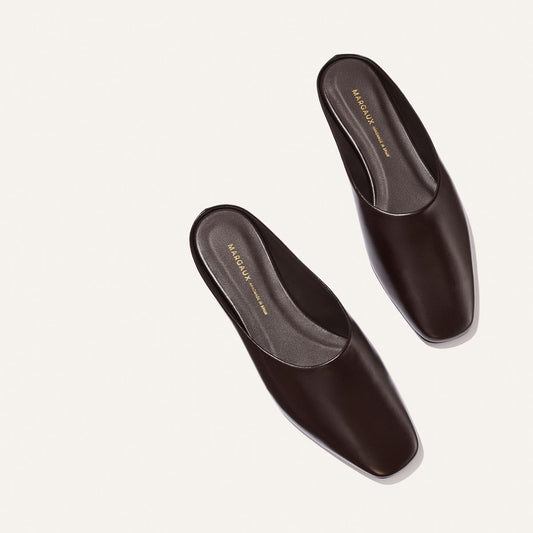 A pair of The Clara - Coffee Calf leather mules by Margaux, featuring a dark brown slip-on design with a square toe, is shown on a light background. The flat, slipper-like silhouette provides effortless everyday style and versatility. Gold Margaux branding appears on the insoles. The shoes are positioned side by side with a slight angle.