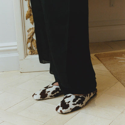 A person in black pants stands on a light wooden floor, wearing Margaux's The Clara - Cow Print Haircalf flats. These comfy slippers have a cow print pattern. Part of a wall and rug are visible in the background.