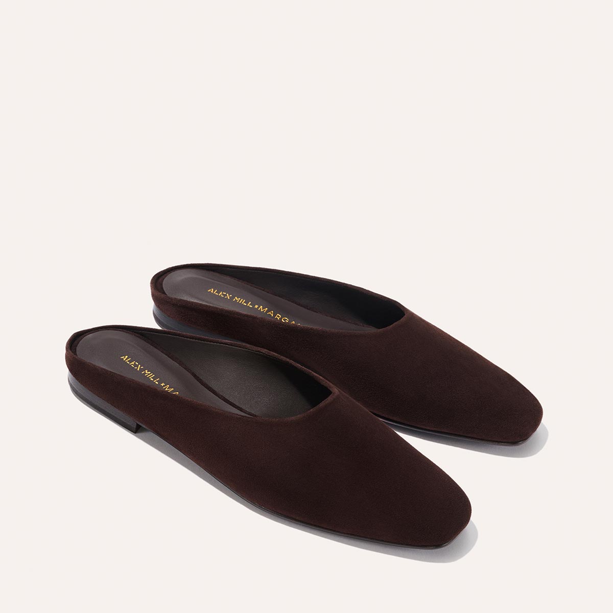The Margaux Alex Mill x Margaux: The Clara - Espresso Suede mules feature a slipper-like silhouette, closed rounded toes, and flat black soles, shown on a light beige background.