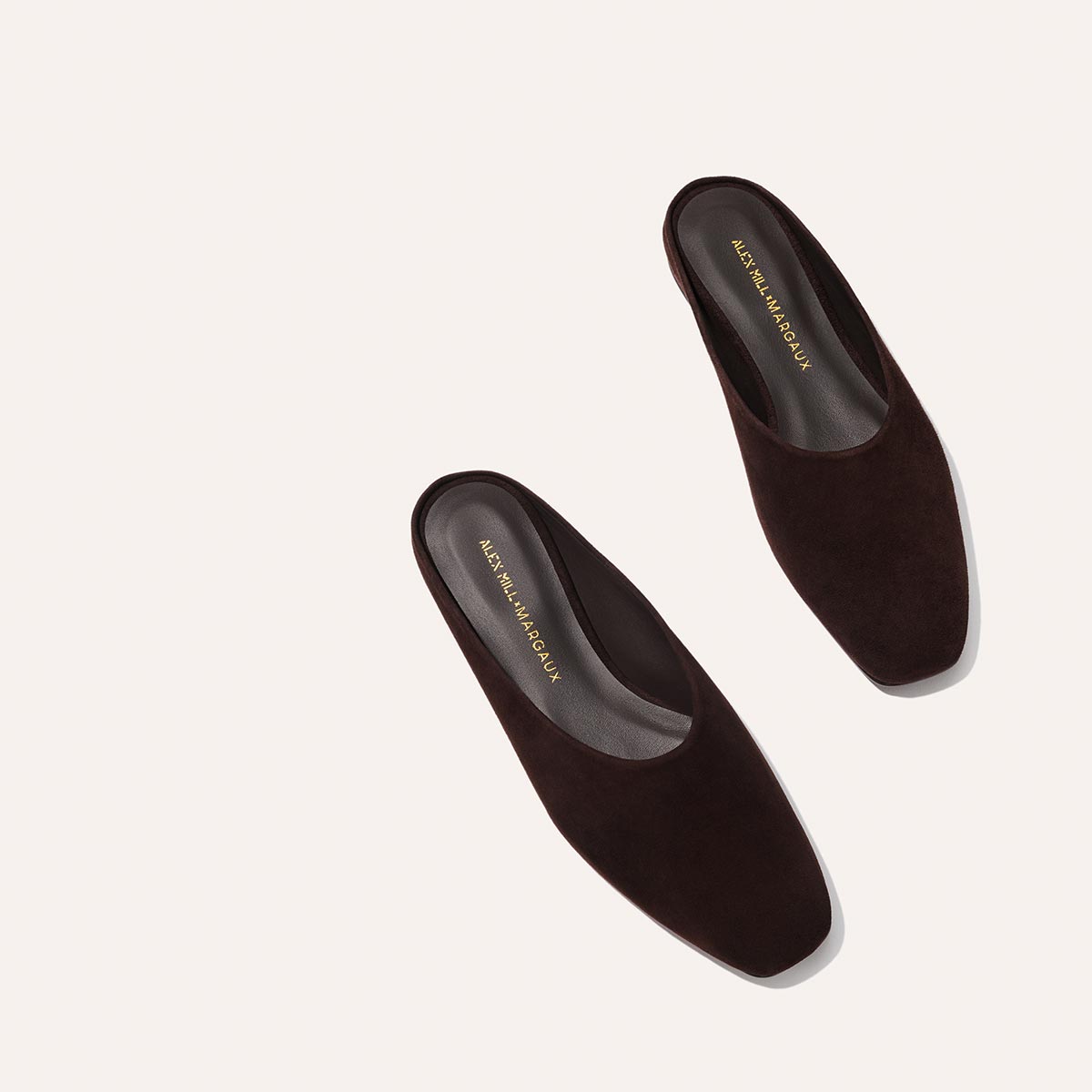 A pair of Alex Mill x Margaux: The Clara - Espresso Suede mules by Margaux, featuring dark brown suede, a flat slipper-like profile, square toes, and gold-stamped insoles, displayed against a light background.