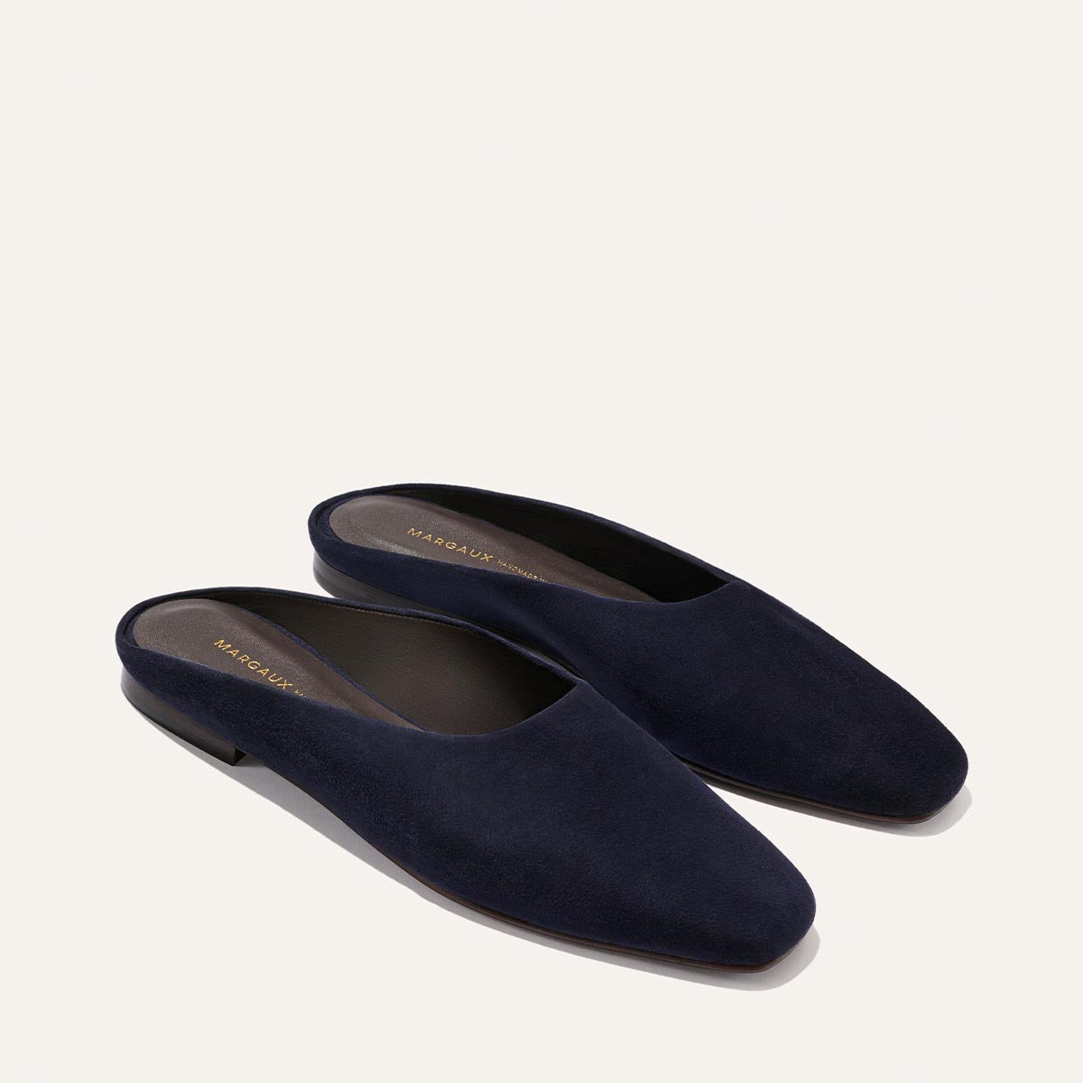 The Clara - Indigo Suede by Margaux: dark navy blue suede slip-on mules with a closed almond toe, flat heel, and slipper-inspired silhouette, shown on a light background.