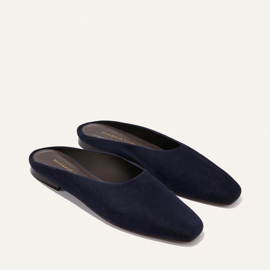 The Clara - Indigo Suede by Margaux: dark navy blue suede slip-on mules with a closed almond toe, flat heel, and slipper-inspired silhouette, shown on a light background.